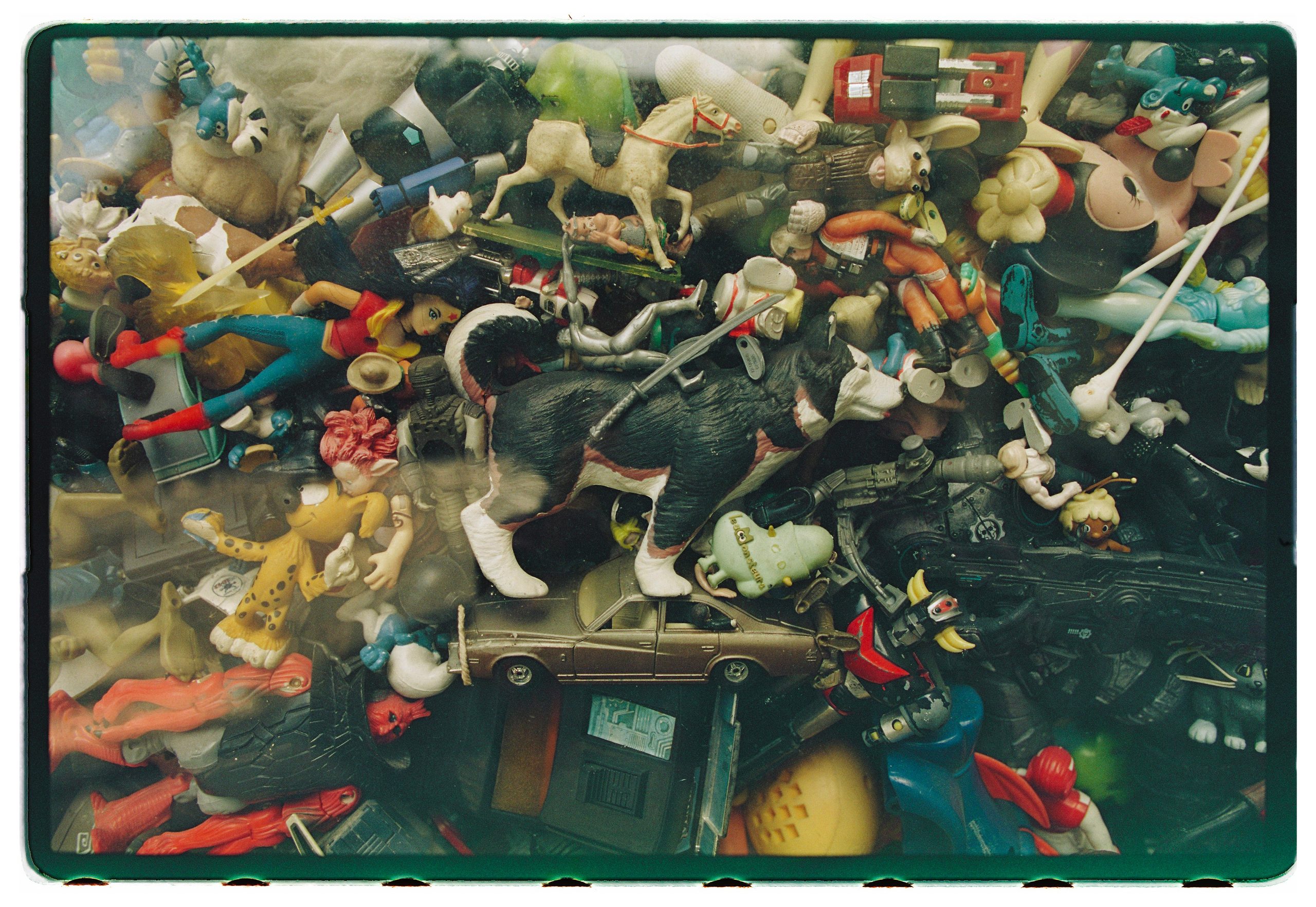 Analog film photograph by Rebeca Lange: a dense case of vintage toy figurines packed behind glass, framed by a green-tinted film border.