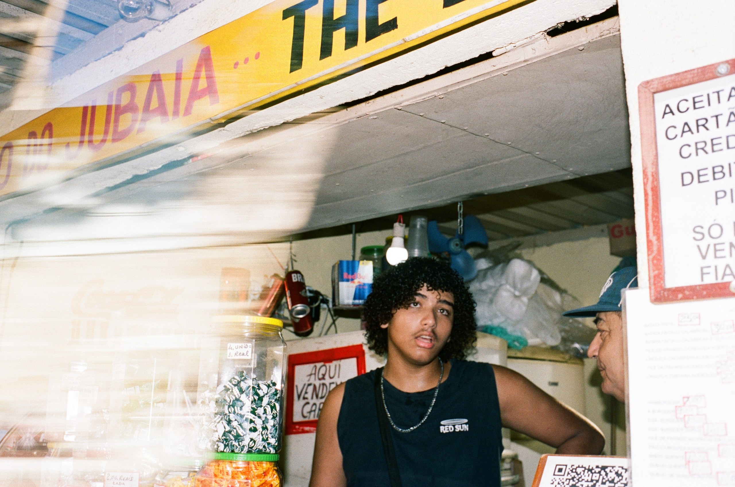 Analog film photograph by Rebeca Lange: a young man behind the counter of a neighbourhood kiosk, warm light leak across the frame.