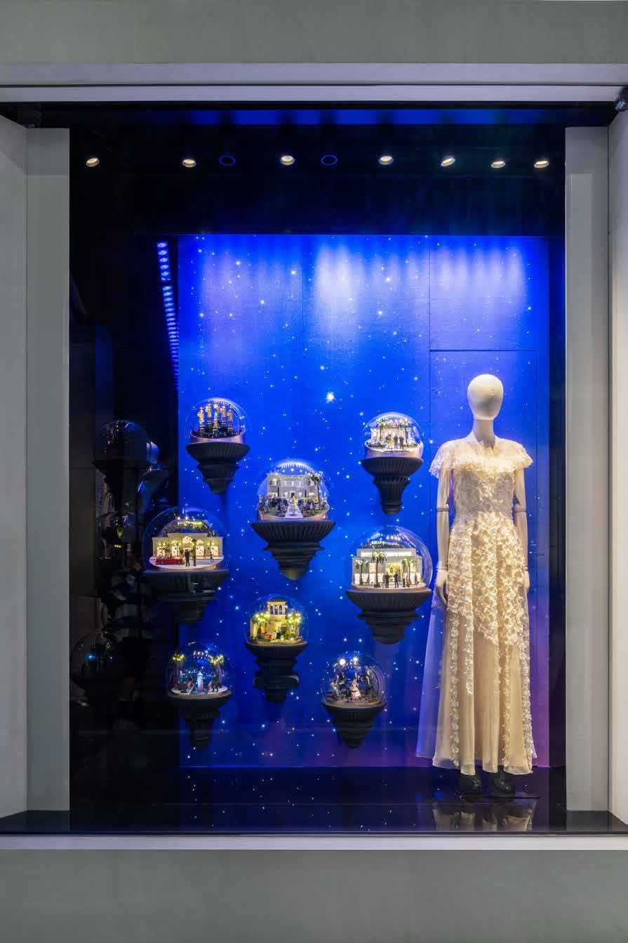 Window display of illuminated snow-globe dioramas beside a white gown on a mannequin.