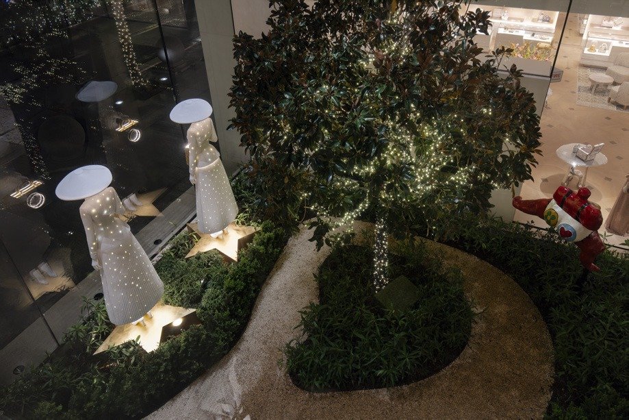 Overhead view of white lantern-dress figures on a lit garden set below a tree.