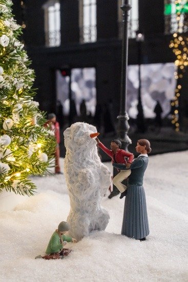 Miniature figures building a snowman beside a lit tree in a snowy diorama.