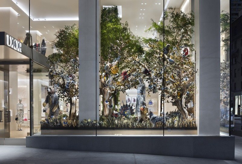 Window scene of leafy trees, birds and figures amid tall grass behind glass.