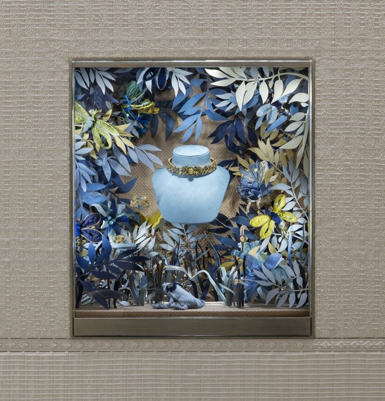 Framed window box with a necklace on a bust amid blue paper leaves and butterflies.