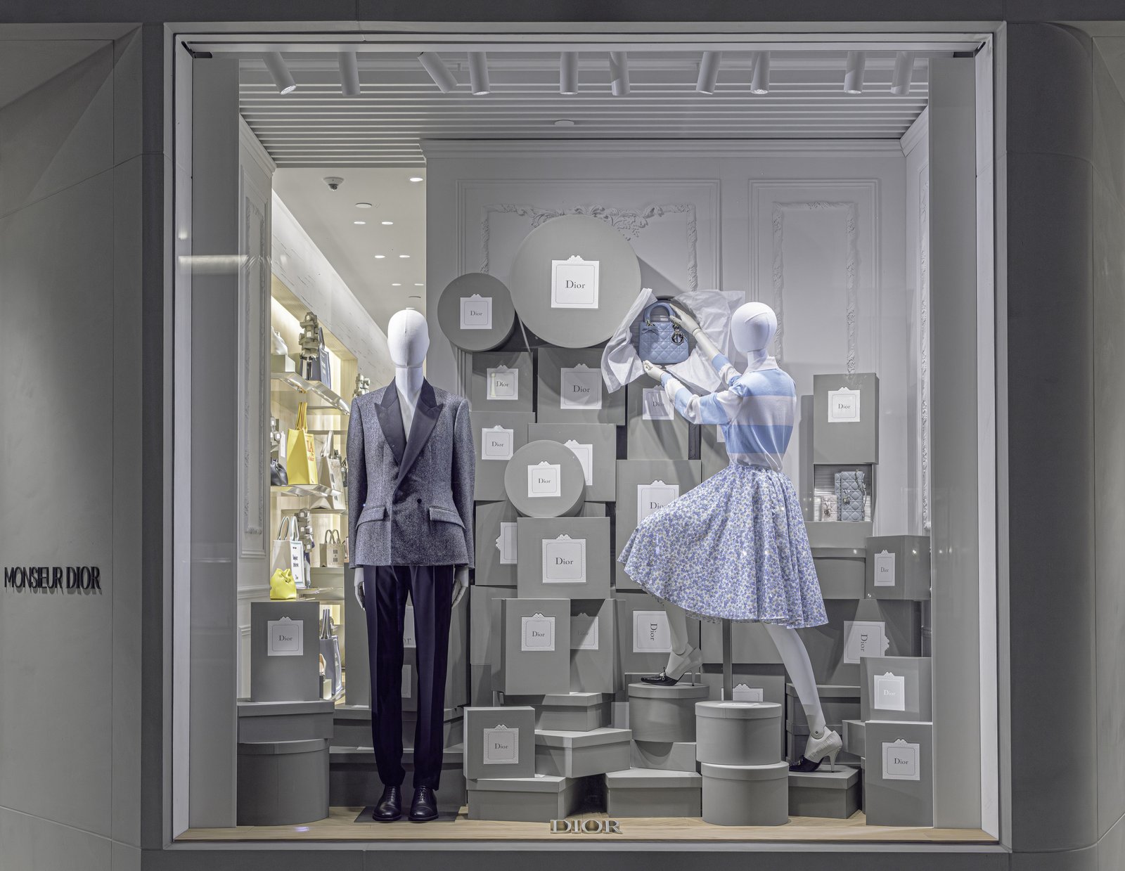 Window display of two mannequins among stacked grey boxes and hatboxes.