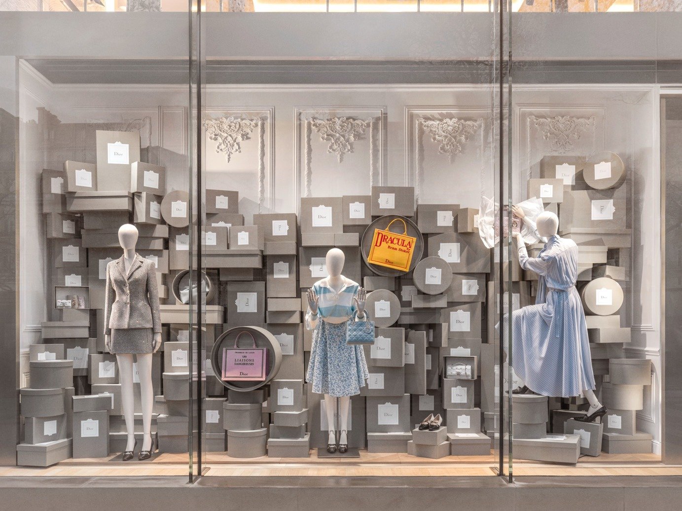 Window display of three mannequins among stacked grey boxes and printed totes.