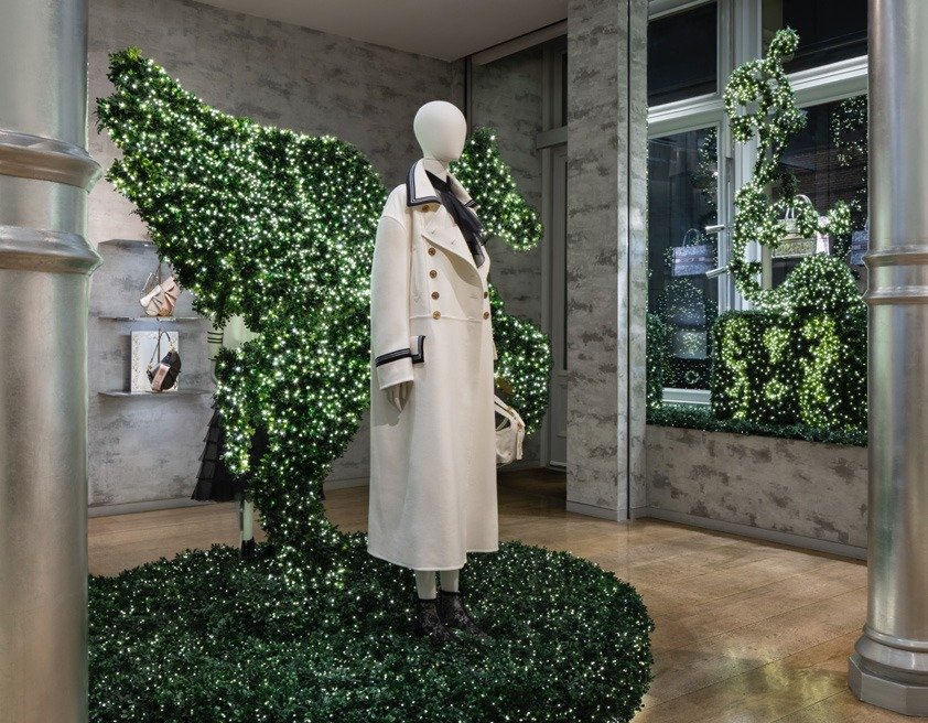 Mannequin in a white coat amid illuminated hedge topiaries in a window display.