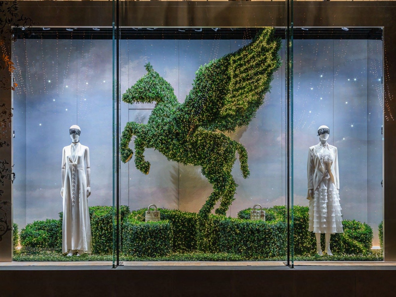 Window display with a foliage-covered winged-horse topiary between two white-clad mannequins.