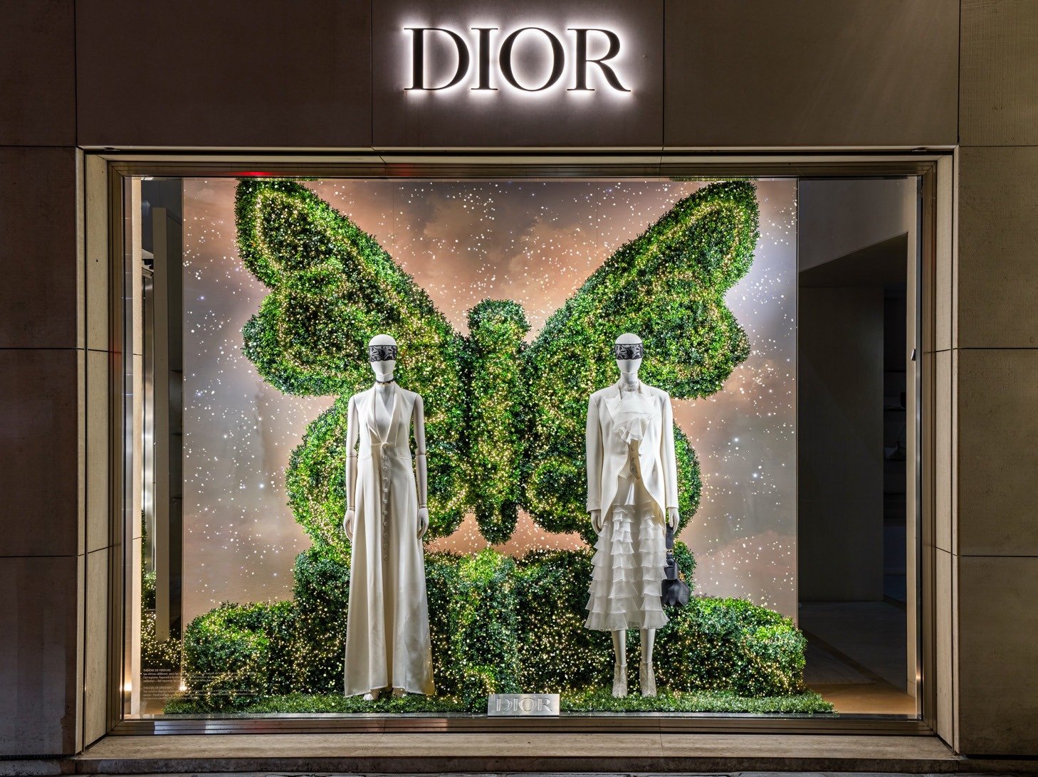 Storefront window with a foliage butterfly topiary behind two mannequins in white.