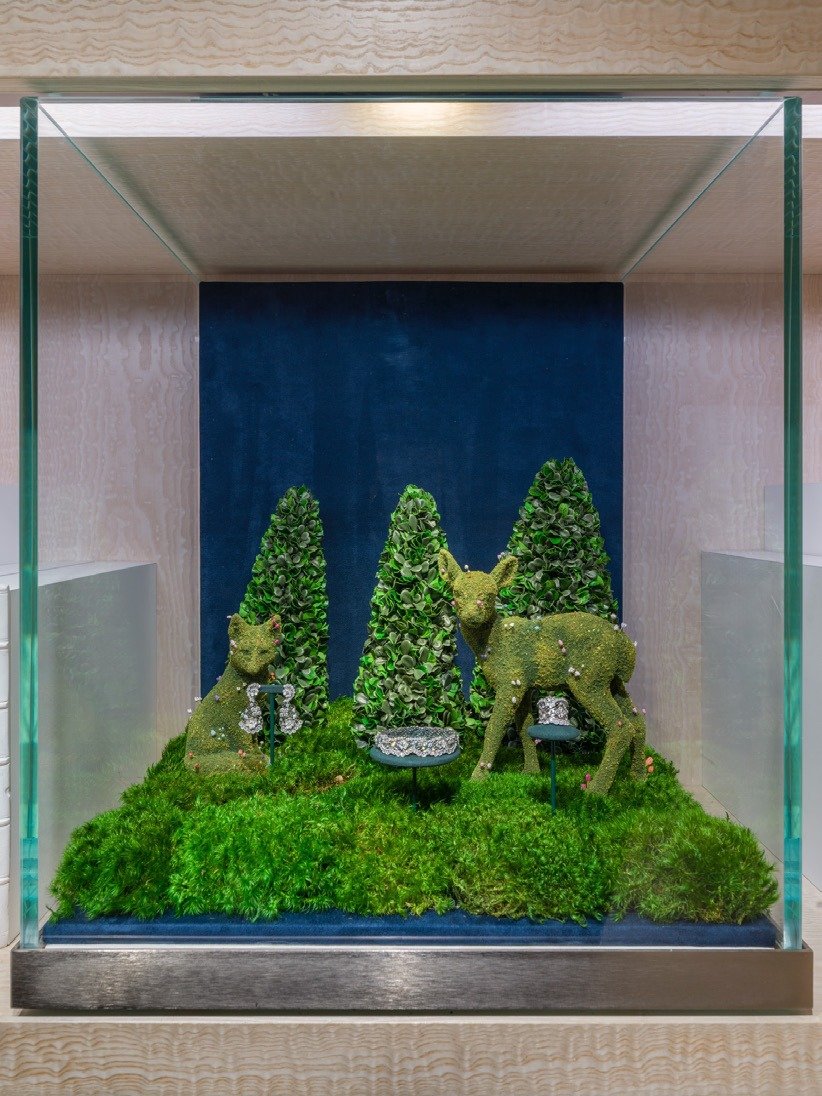 Glass case holding moss deer figures and topiary trees with jewellery on a blue ground.