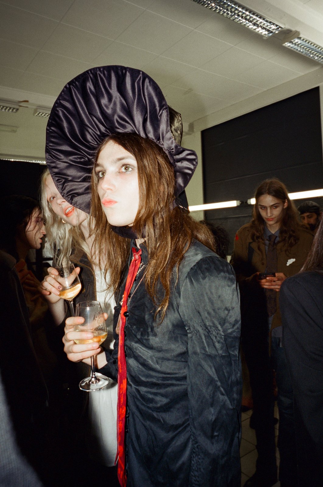 A model in a dramatic oversized black satin hat holds a glass of white wine at a dim indoor gathering, shot on flash film.