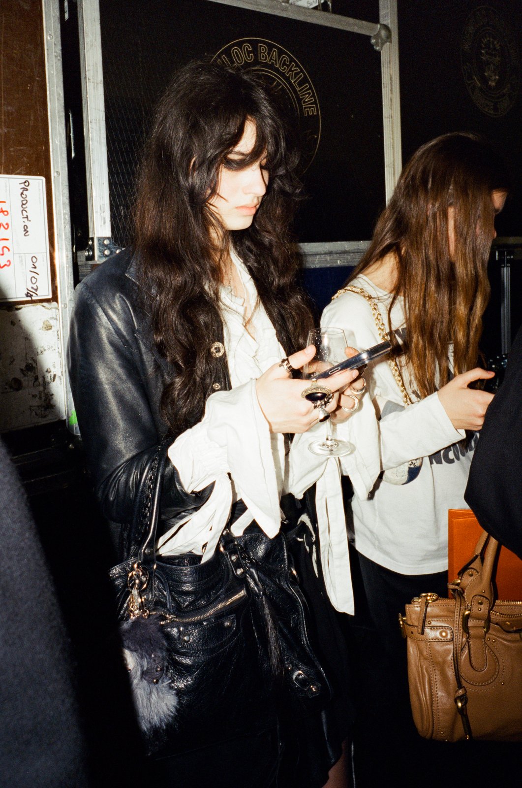 A dark-haired person in a black leather jacket checks a phone while holding a wine glass, with a tan handbag nearby, backstage flash photo.