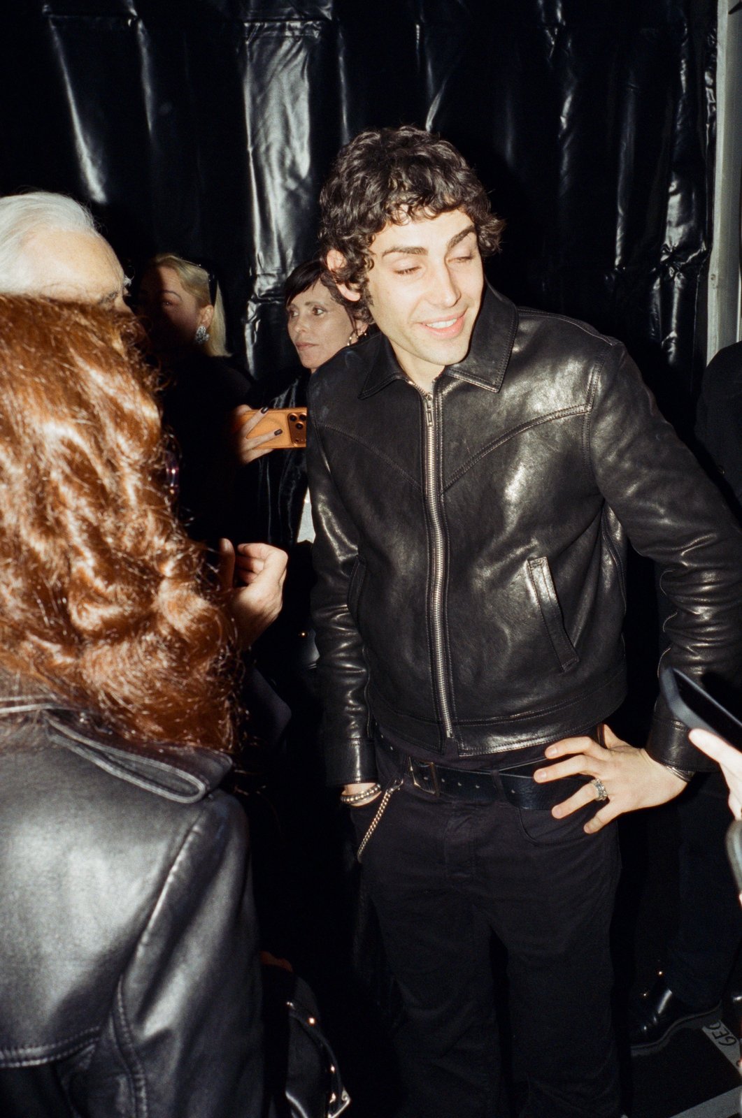 A smiling young man in a black leather jacket stands among people in a dark room, warm film grain.
