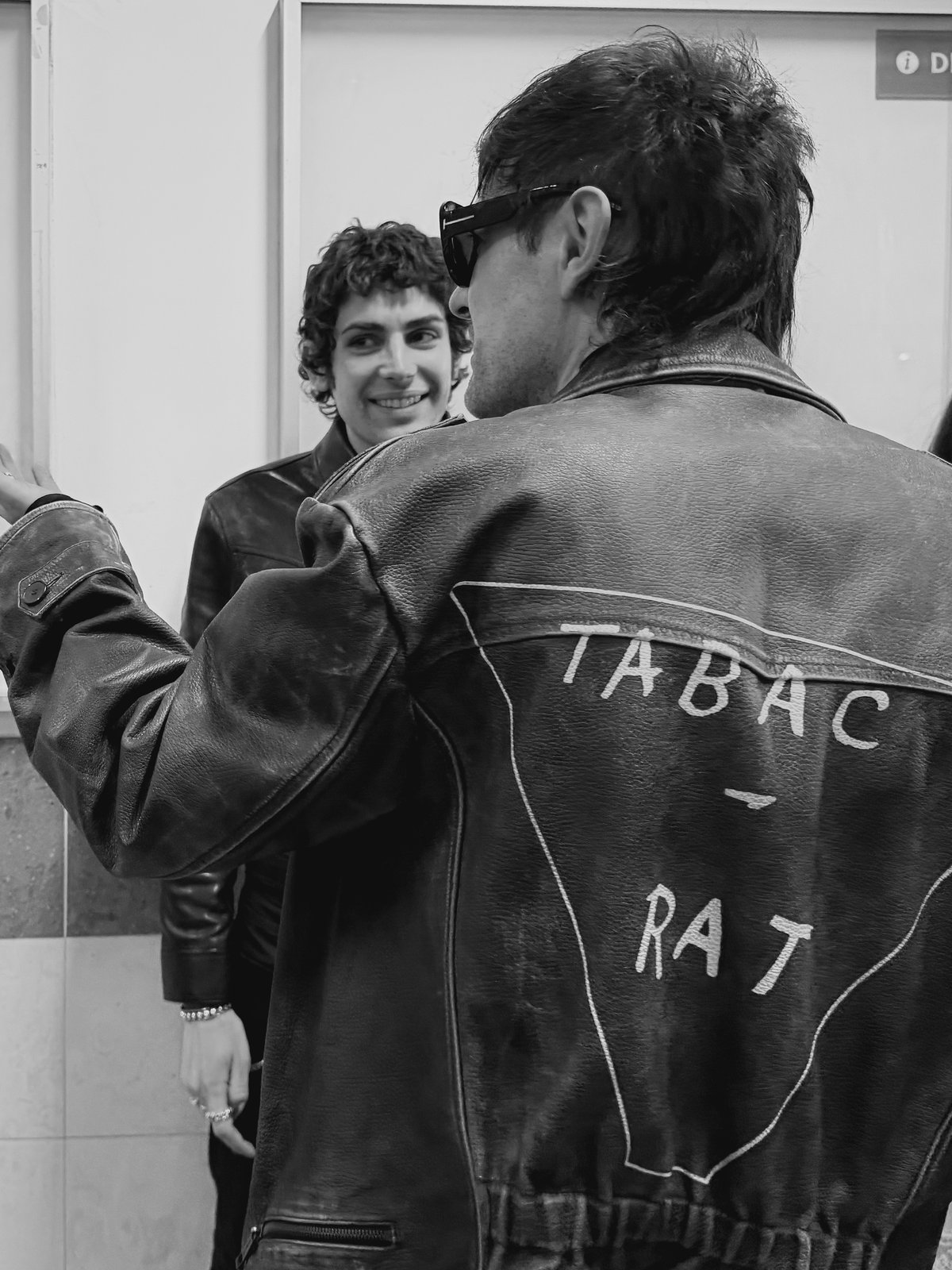 Black-and-white shot of a man in sunglasses and a hand-lettered leather jacket facing a smiling curly-haired man.