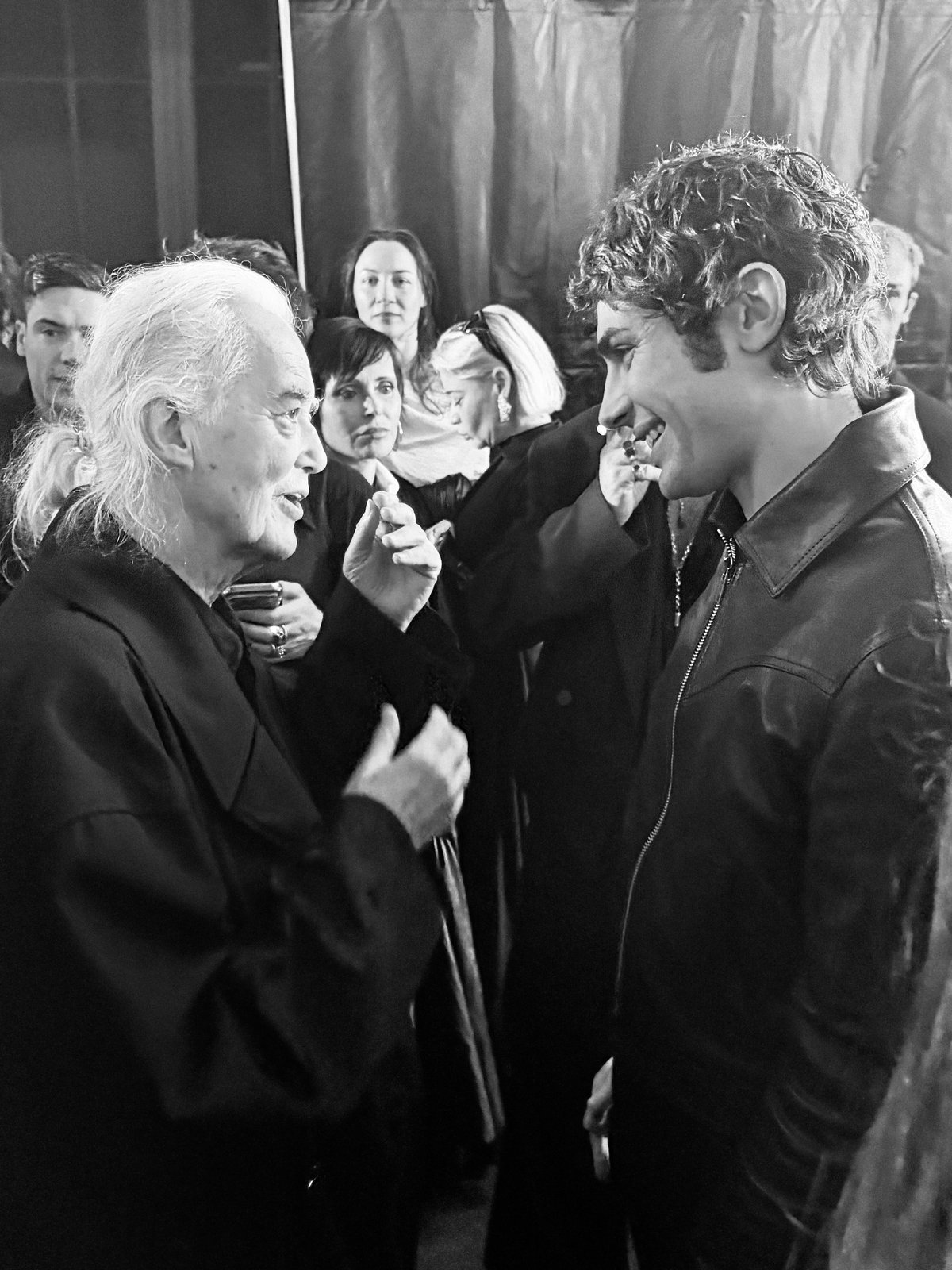 Black-and-white photo of a smiling curly-haired young man in leather talking with an older long-haired man in black, others behind.