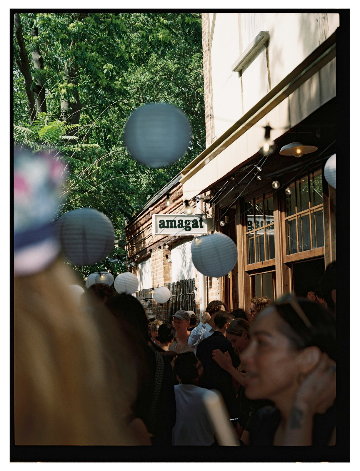 Paper lanterns hang over a crowd outside a restaurant with a lit sign, backed by trees.