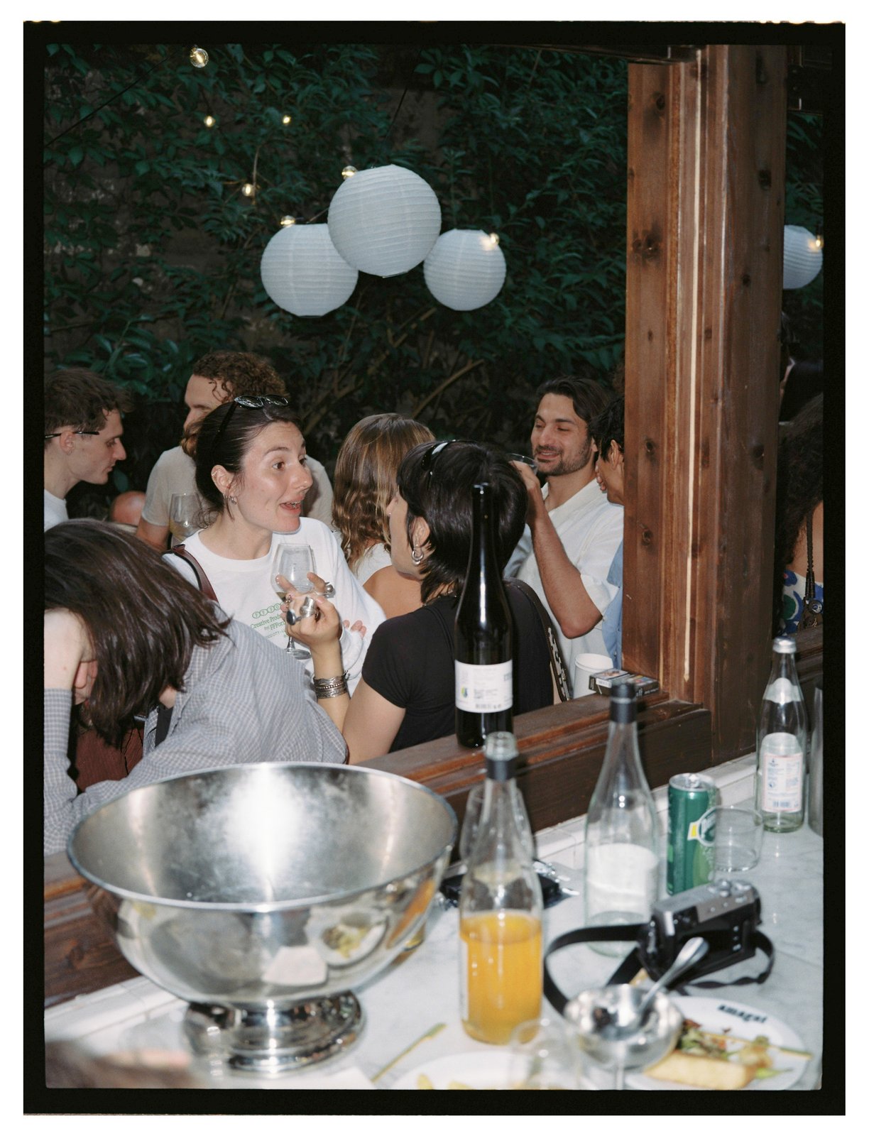 Guests mingle with wine glasses at a bar with bottles and a punch bowl under paper lanterns.