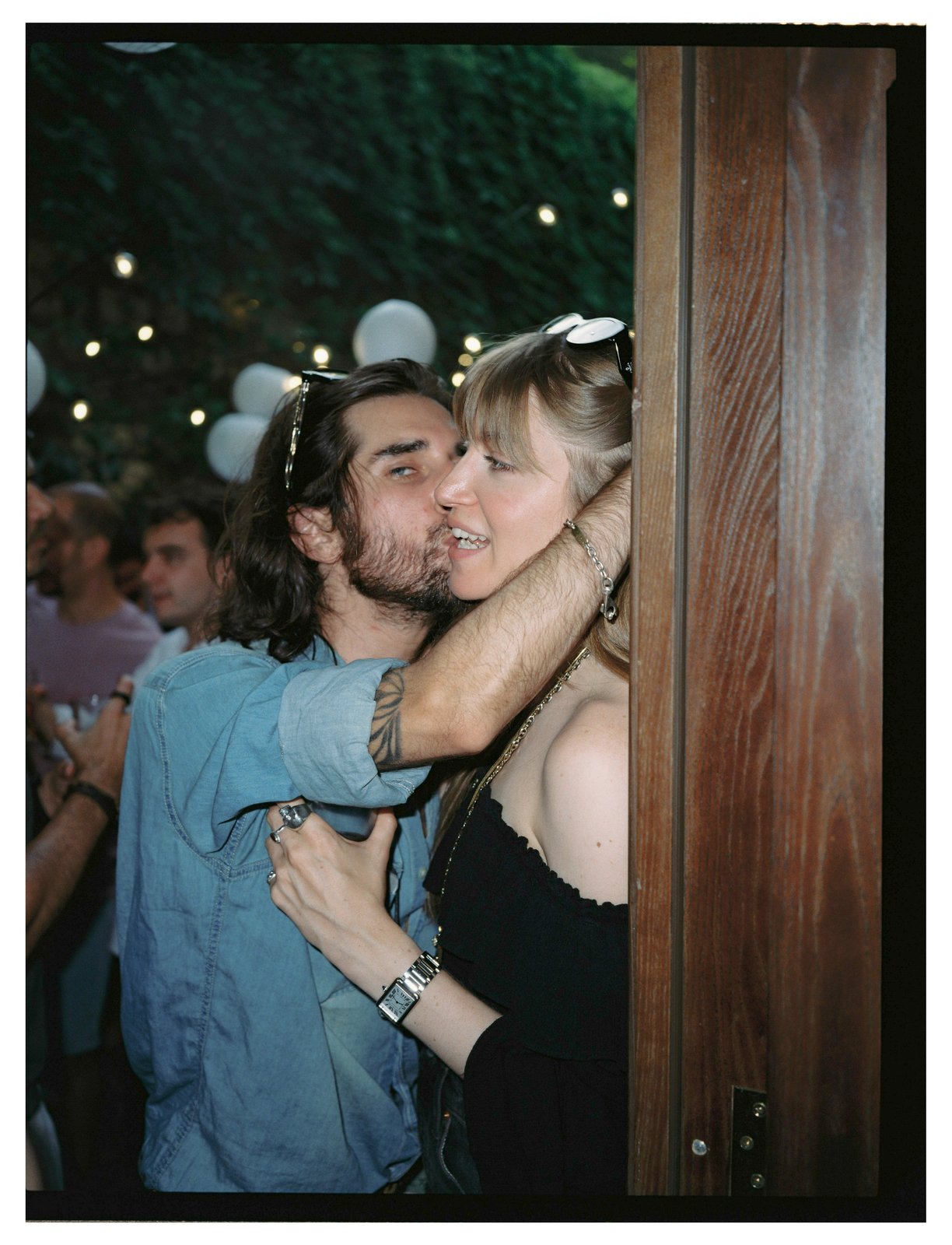 A man in a denim shirt embraces a blonde woman at a night party strung with lights.