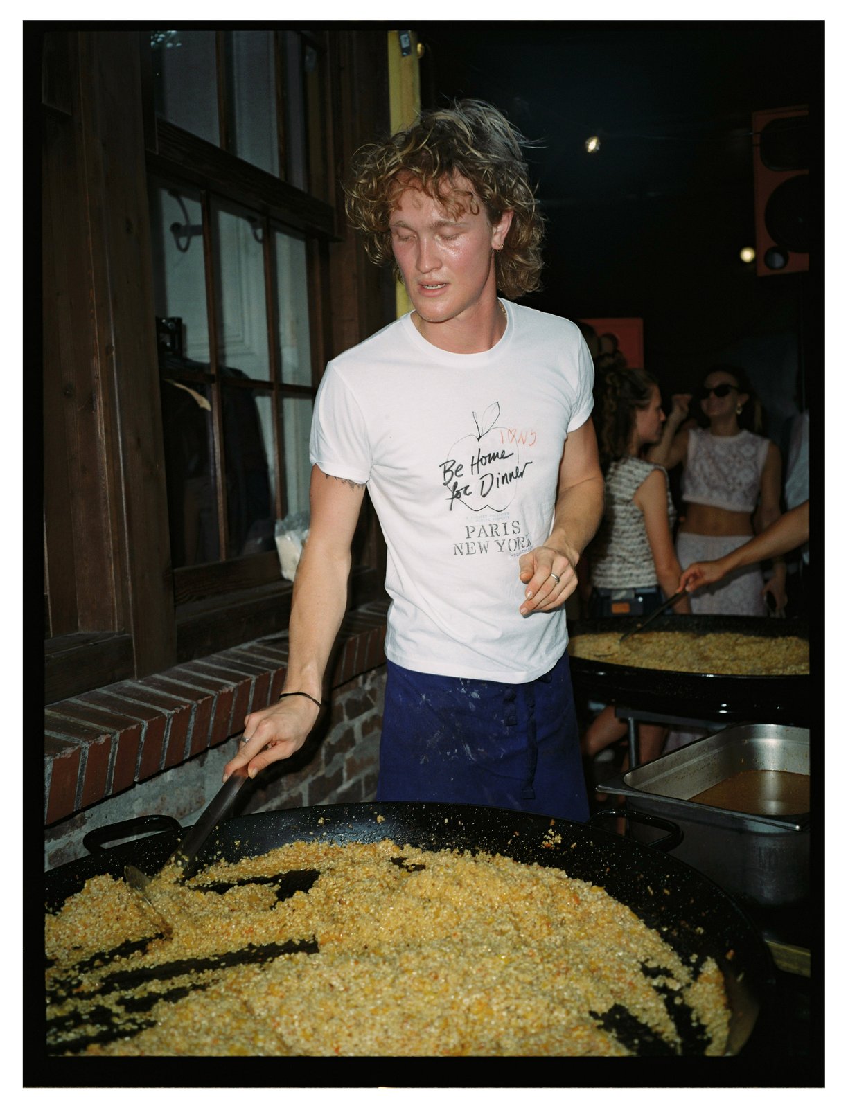 A man in a printed tee stirs a large pan of rice at an evening event.