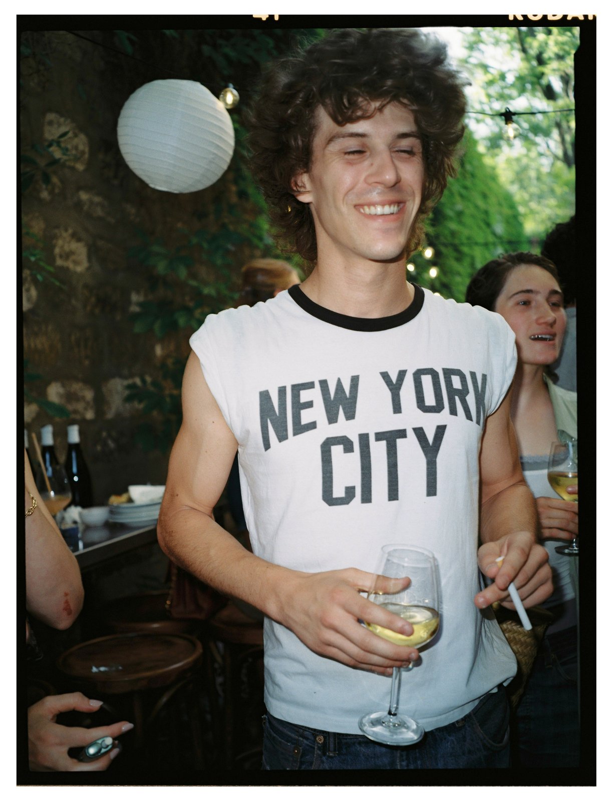 A smiling young man in a graphic shirt holds a glass of white wine at a garden party.