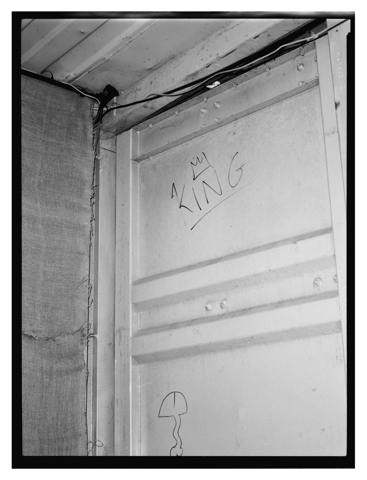 Black-and-white flash frame of a corrugated wall with hand-drawn crowned graffiti and cabling overhead.