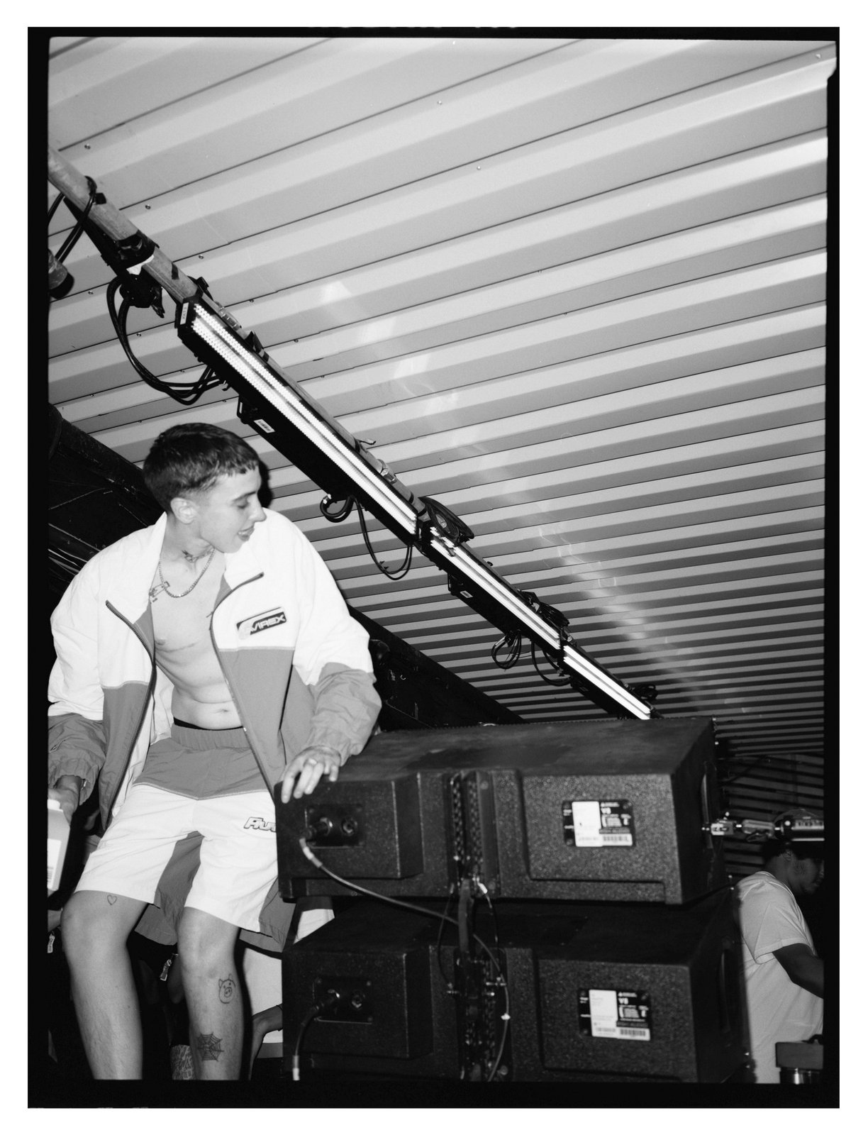 Black-and-white shot of a person in a white jacket and shorts leaning on stacked speaker cabinets under a metal roof.