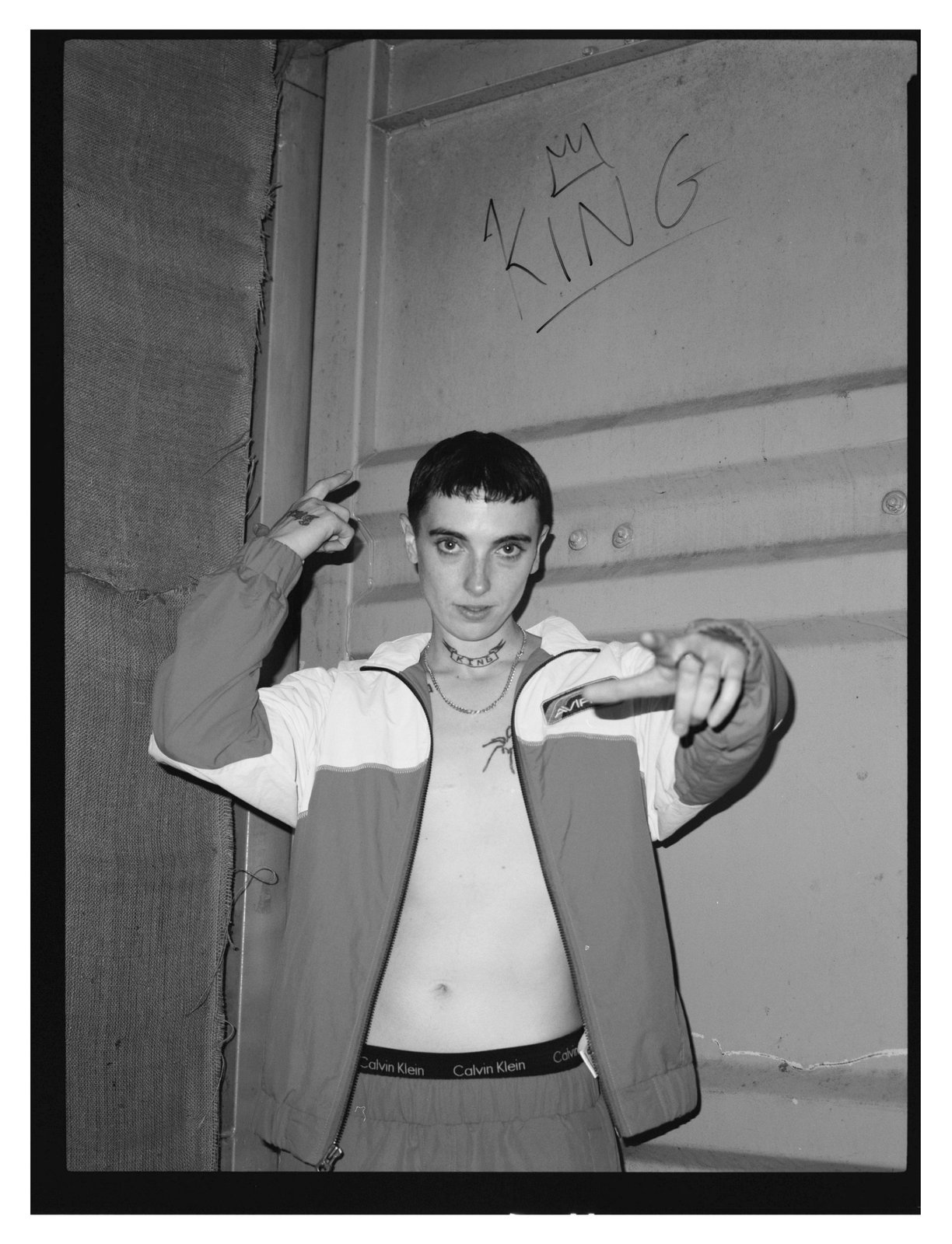 Black-and-white portrait of a young person in an open zip jacket gesturing beneath crowned graffiti.