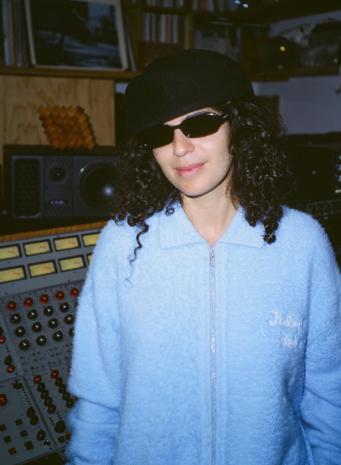 A curly-haired person in sunglasses, a pale-blue fleece jacket and cap stands in a recording studio by a mixing desk.