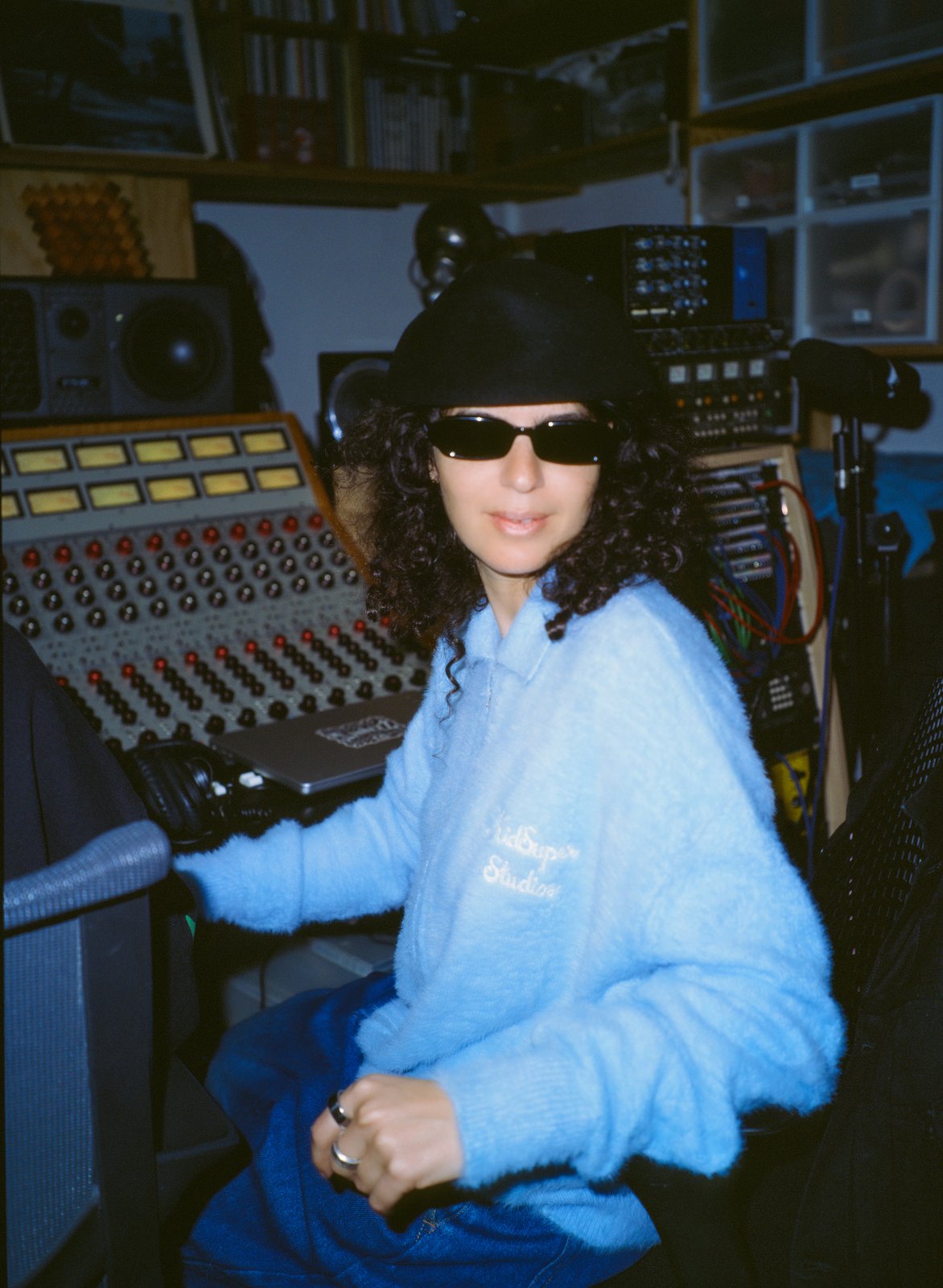 A person in sunglasses, black cap and fluffy blue jacket sits amid studio mixing consoles and equipment.