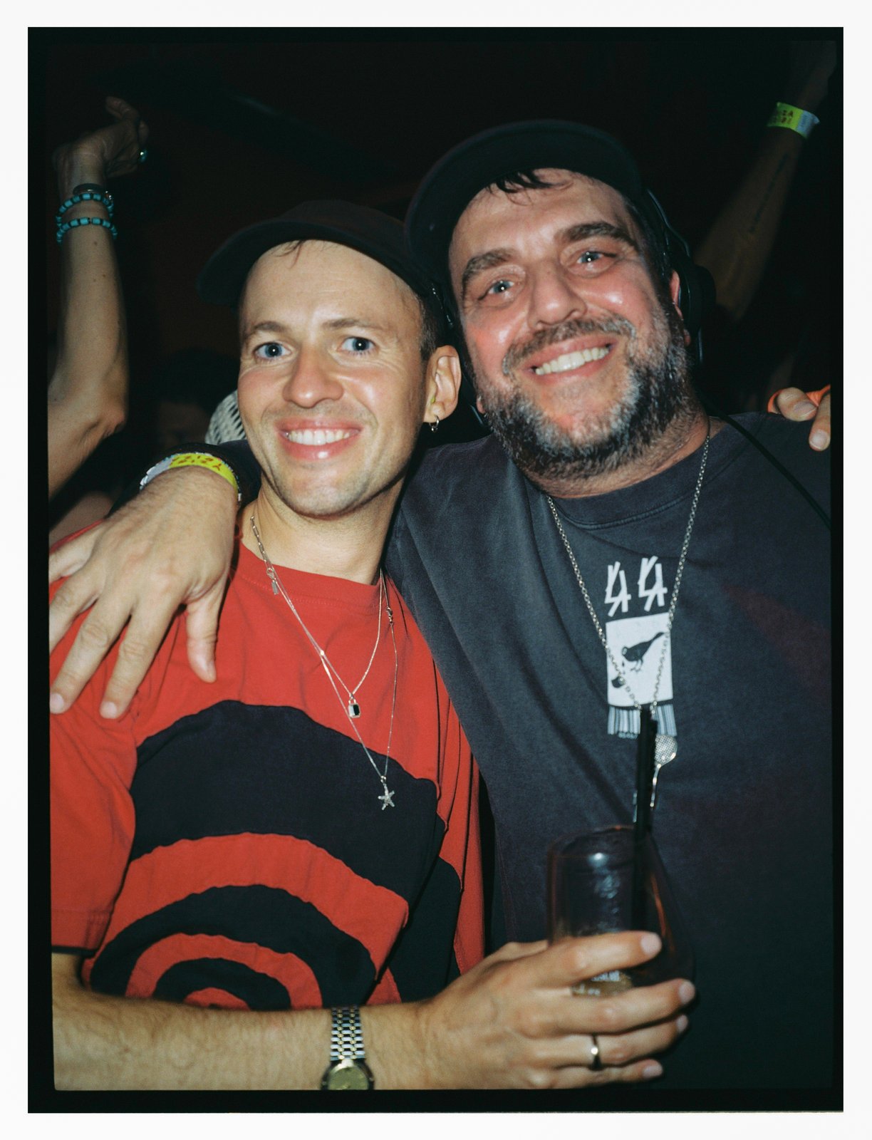 Two smiling men in caps embrace at a party, one in a red top, flash-lit analog frame.