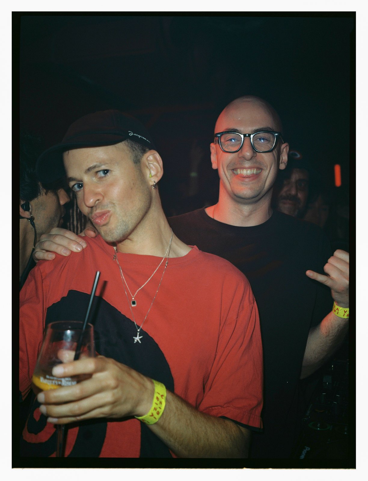 Two grinning men at a dark club, one in a red shirt holding a drink, bright flash.