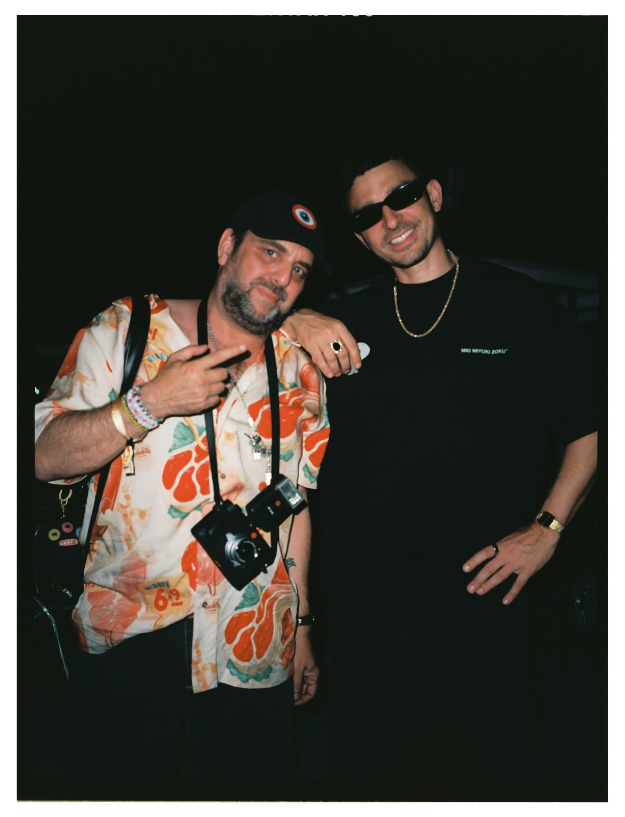 Night flash portrait of two men posing close; one in a cap and orange-print shirt with a film camera, the other in black with sunglasses and a gold chain.