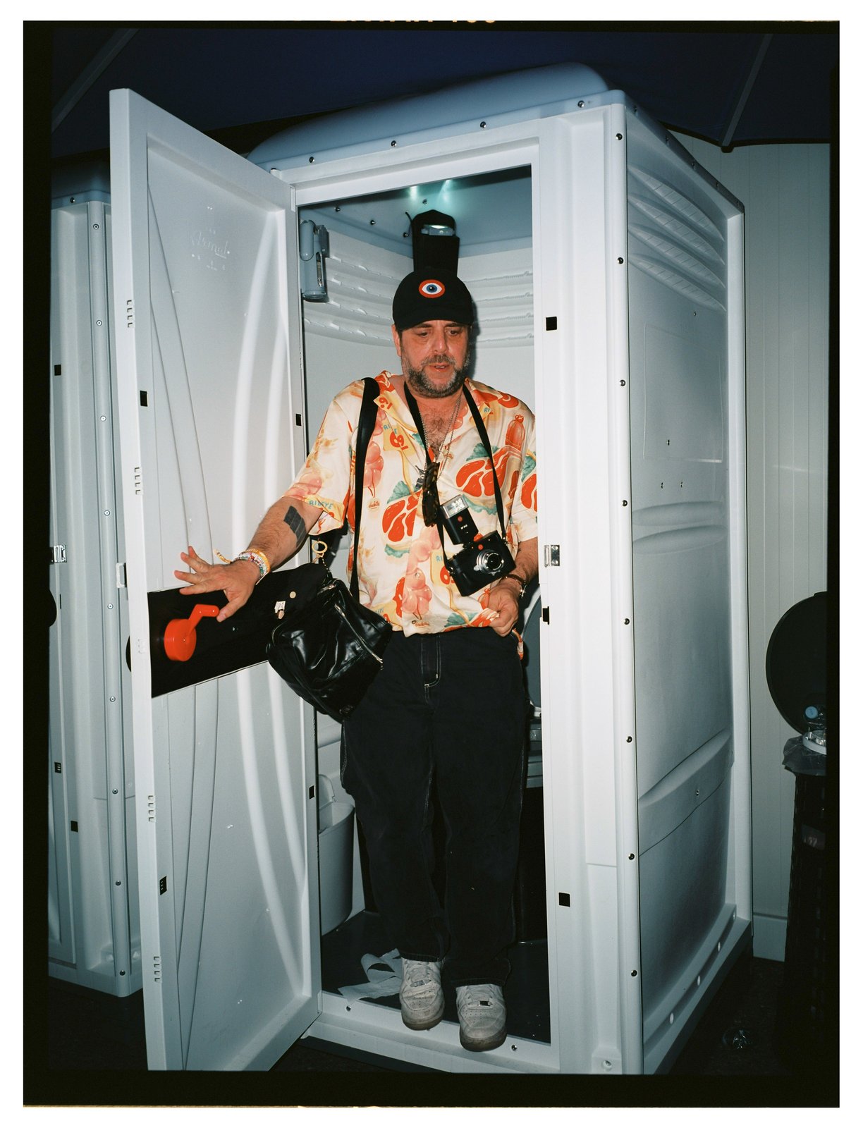 A man in a cap and orange-print shirt, film camera round his neck, stepping out of a white portable toilet at night.