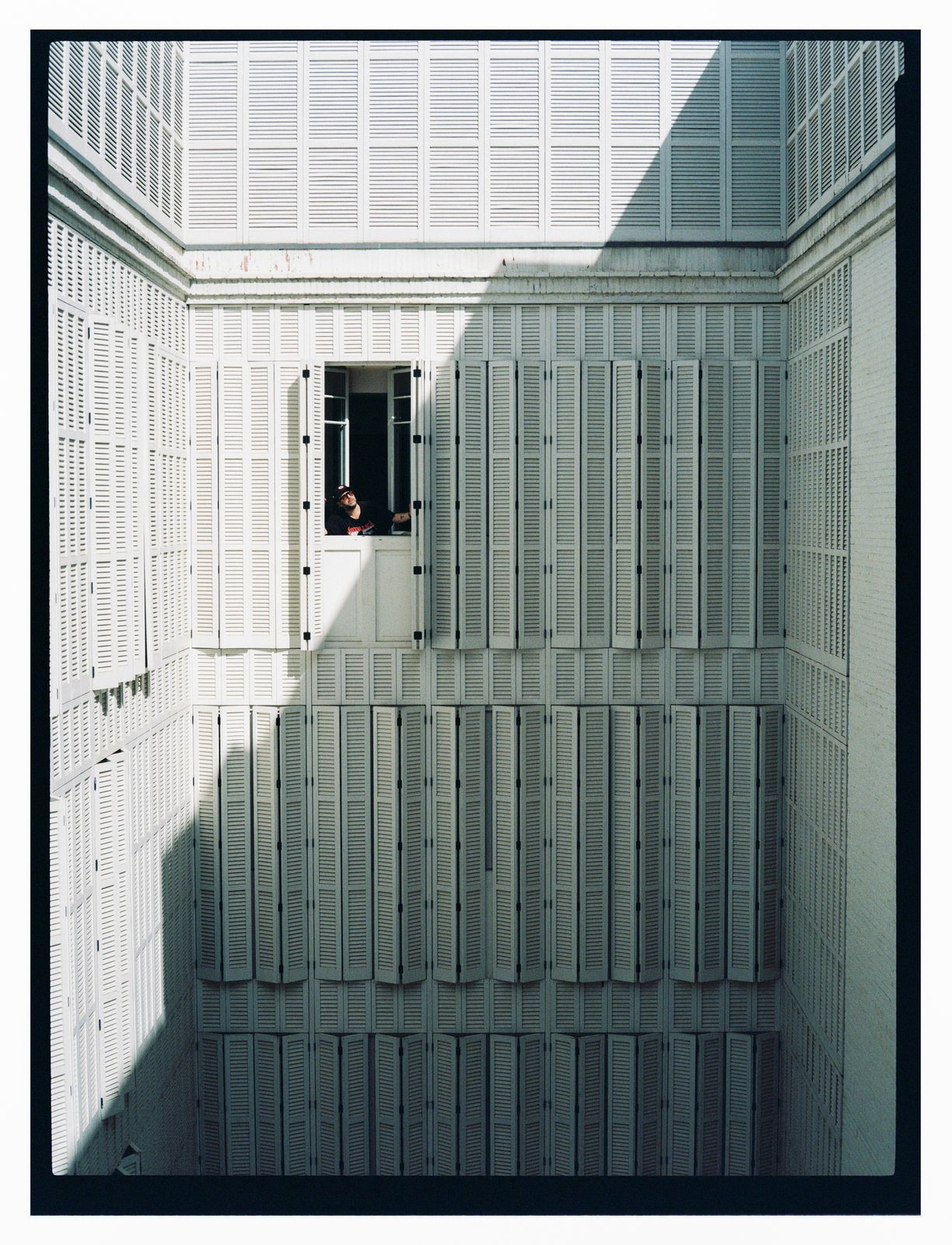 Daylight shot of a small figure in a cap leaning from an open window amid a vast grid of white louvred shutters.
