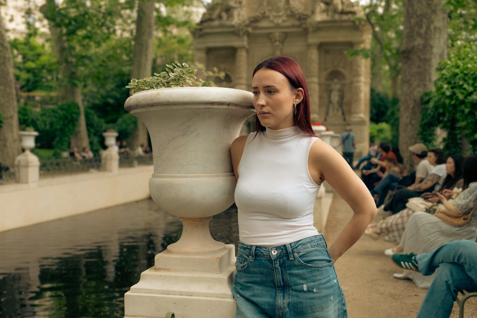 A red-haired woman in a white top and jeans stands beside a stone urn in a park.