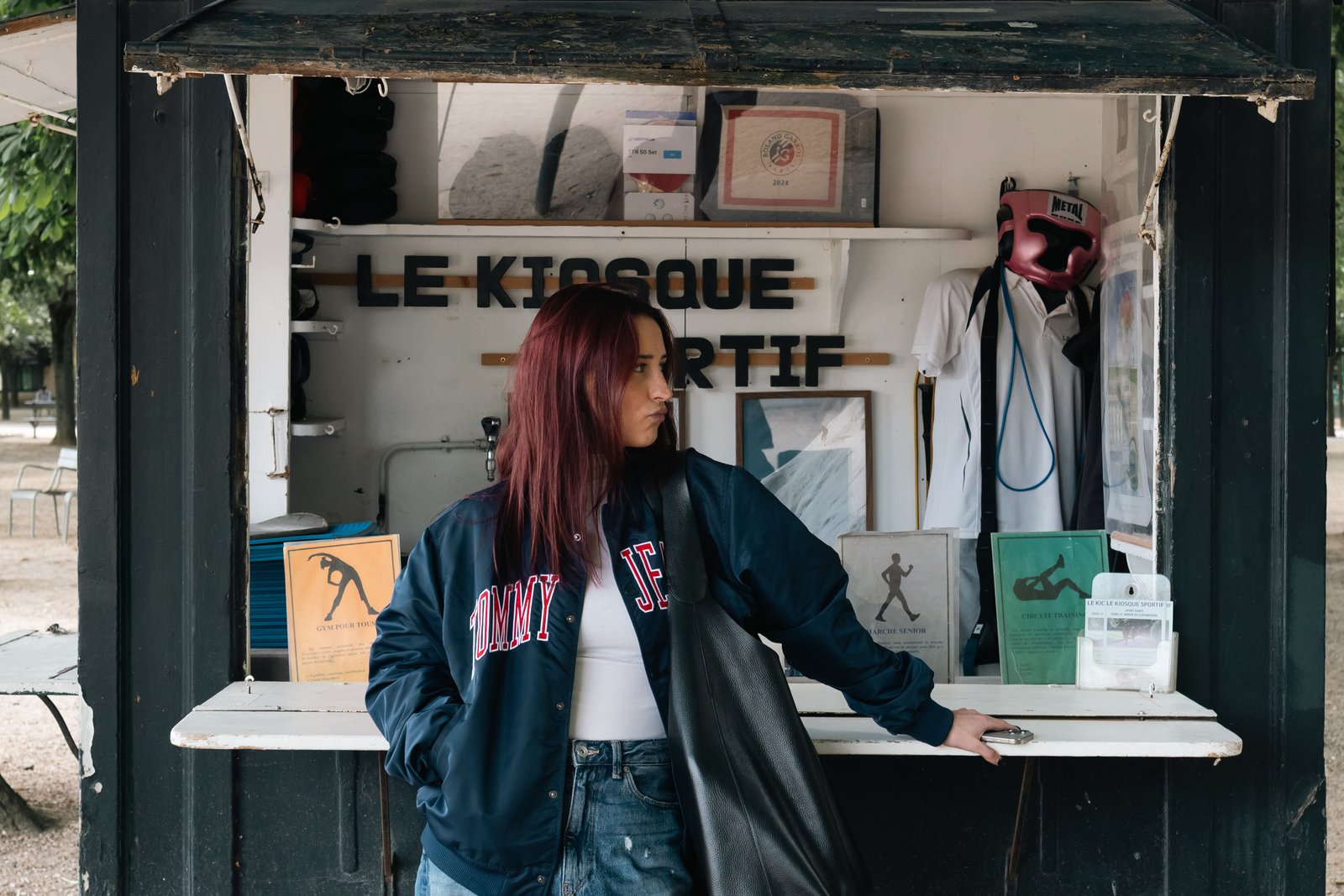 A red-haired woman in a navy varsity jacket leans against a park kiosk.