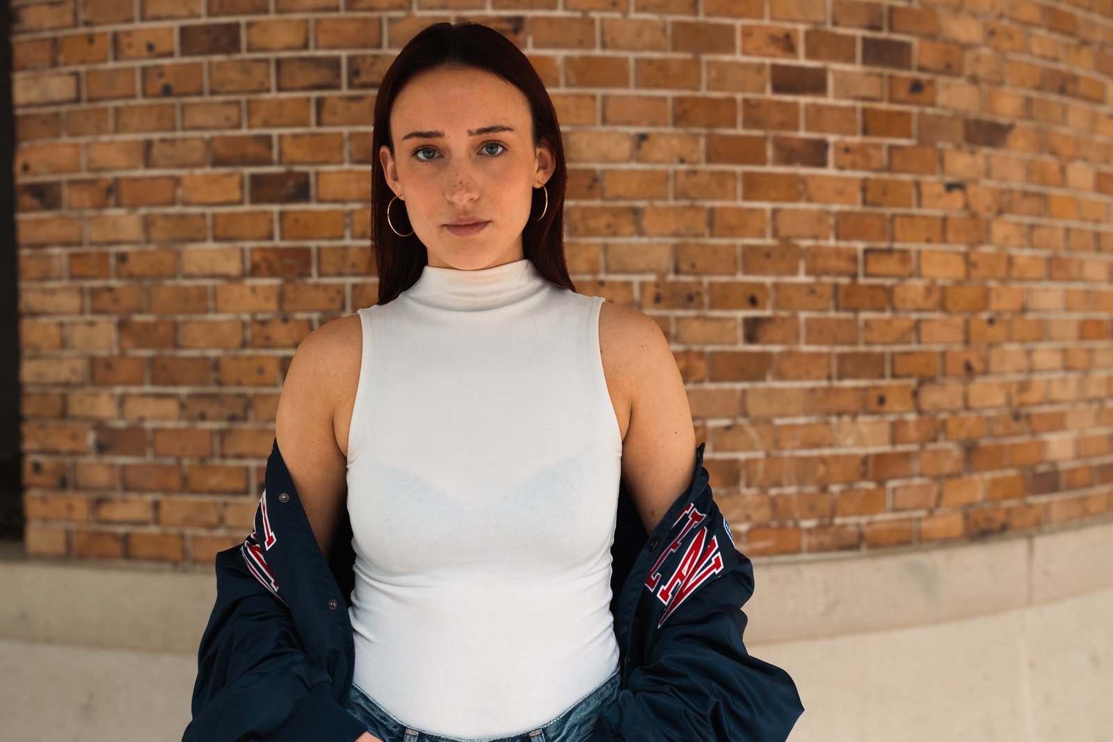 A red-haired woman in a white top and navy varsity jacket stands before a curved brick wall.