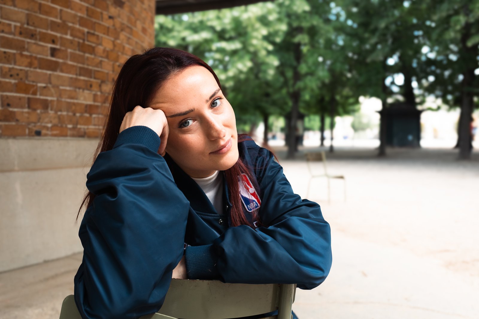 A red-haired woman in a navy jacket rests her head on her hand at an outdoor table among trees.