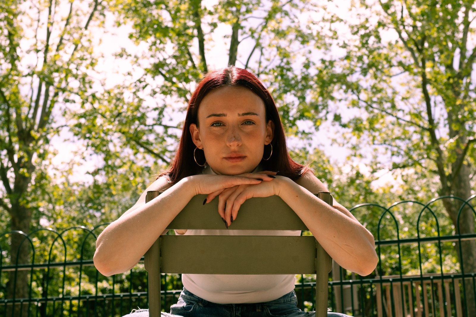 A red-haired woman leans on the back of a chair in a sunlit park with green foliage.