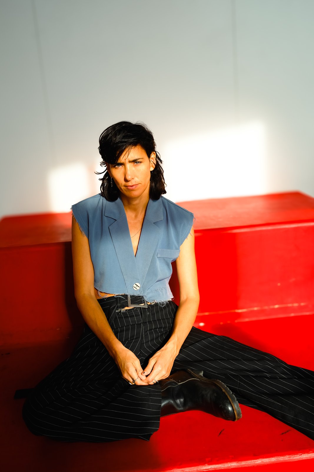 A person in a blue sleeveless top and pinstripe trousers sits on red steps against a white wall.