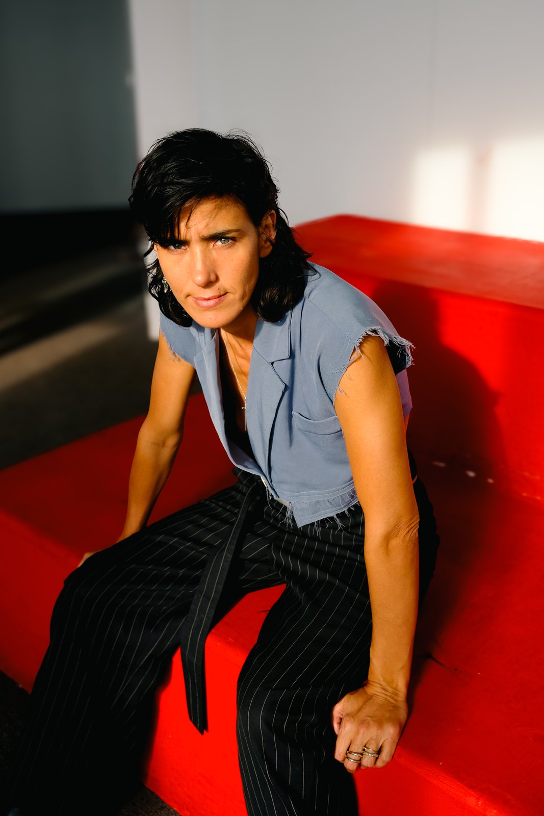 A person in a blue cropped vest and pinstripe trousers sits on red steps in raking light.