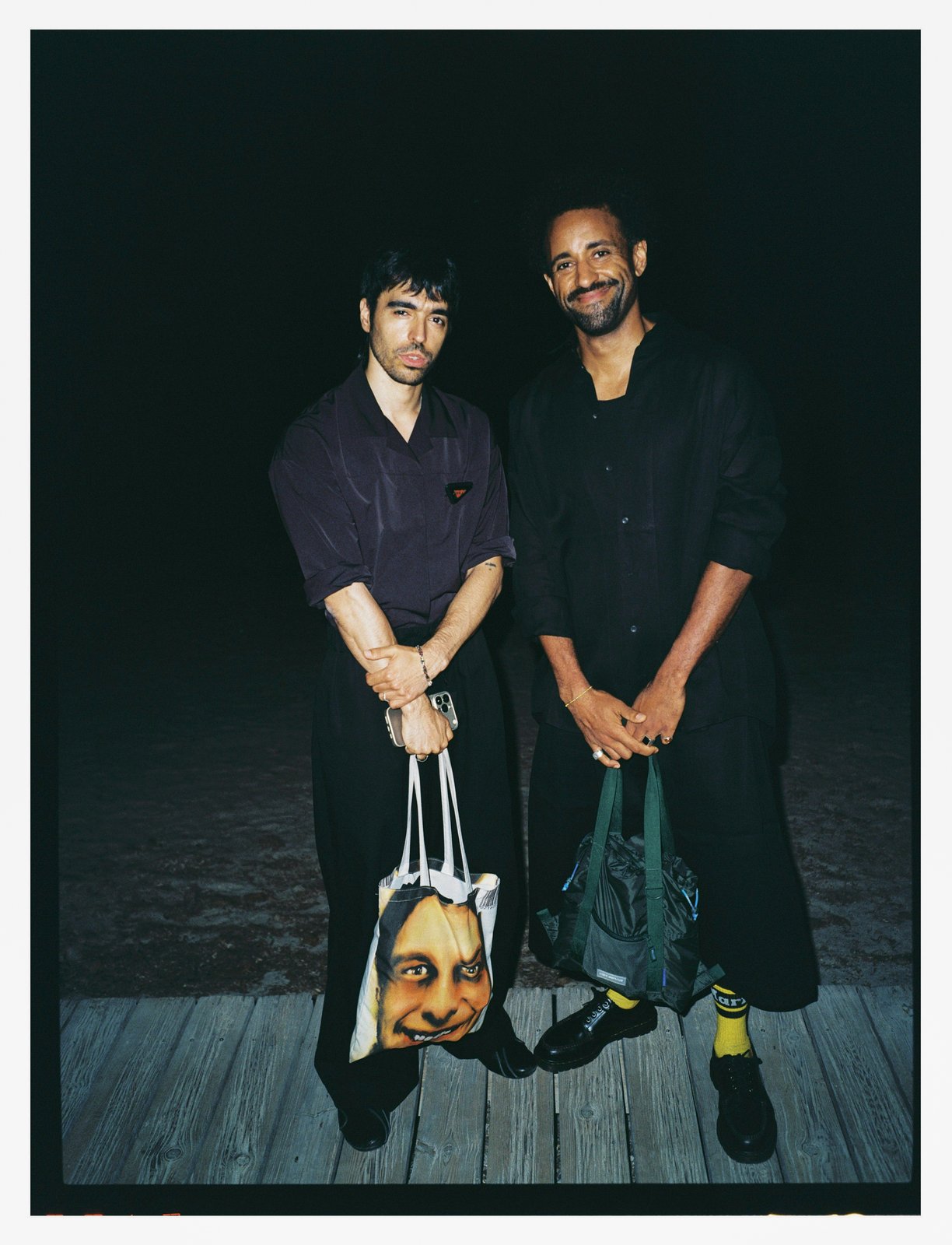 Night flash portrait of two men in dark clothes on a boardwalk holding tote bags, one printed with a screaming face.