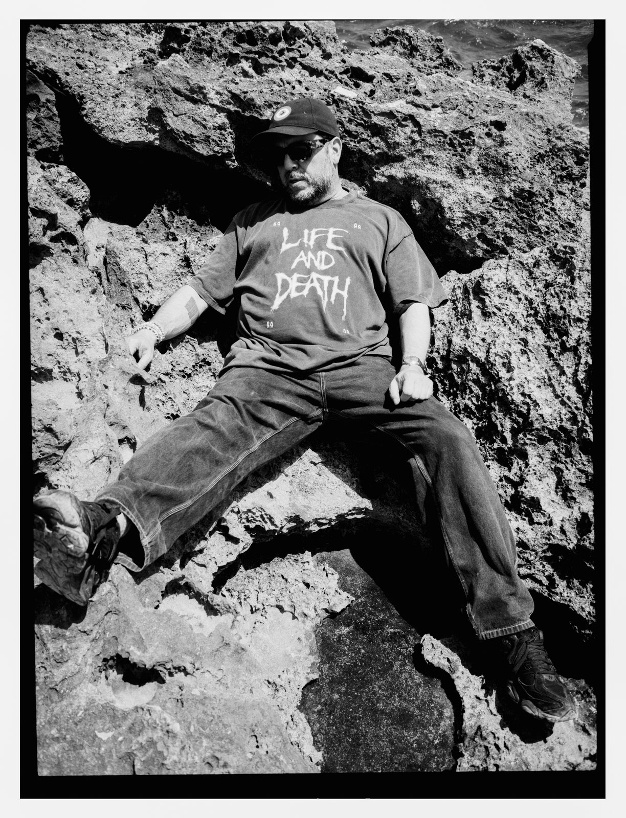 Black-and-white portrait of a bearded man in a cap and sunglasses reclining across jagged coastal rocks in a "Life and Death" T-shirt.
