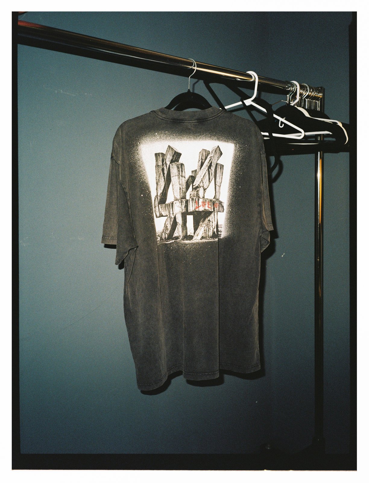 A faded black T-shirt printed with a photographic graphic hangs on a rail against a blue wall, spare hangers beside it.