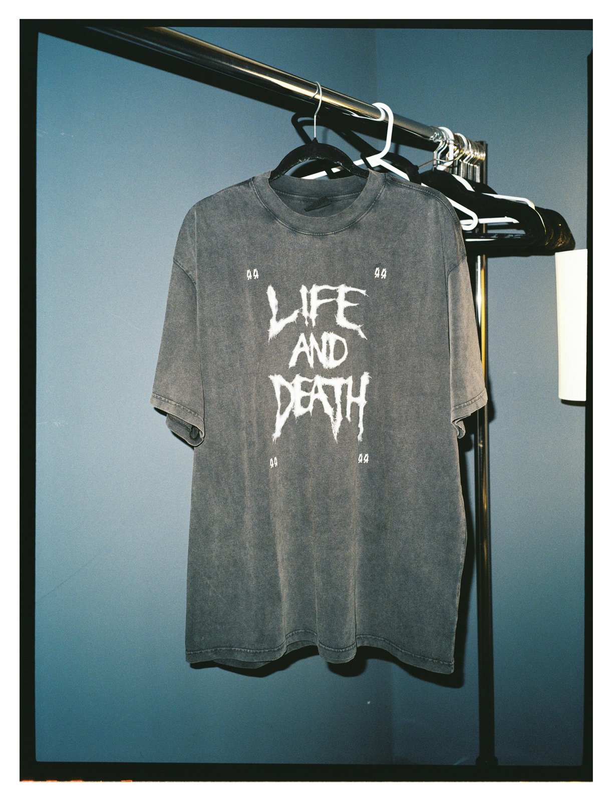 A washed-grey T-shirt with spiky "Life and Death" lettering hangs on a clothes rail against a blue-lit wall.