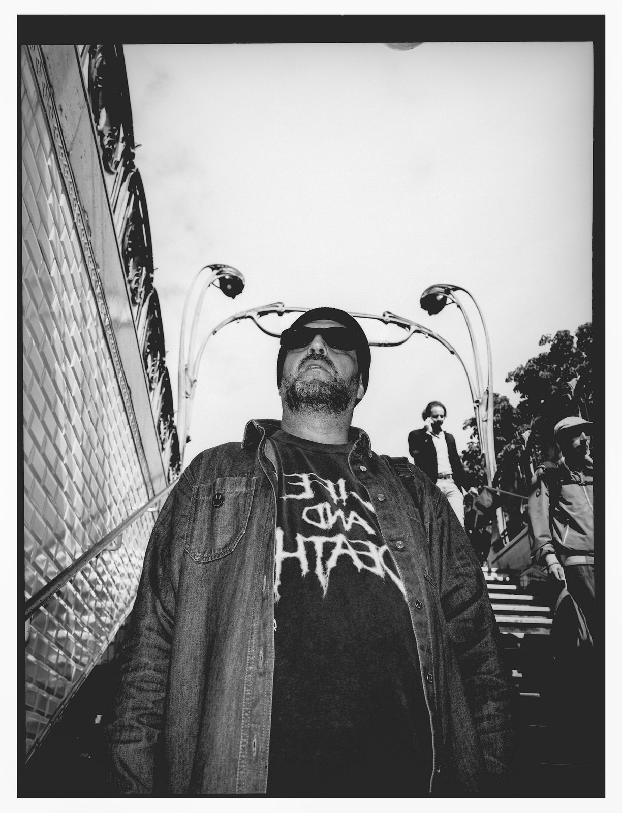 Low-angle black-and-white shot of a bearded man in sunglasses and a denim jacket emerging from a metro staircase, figures behind.