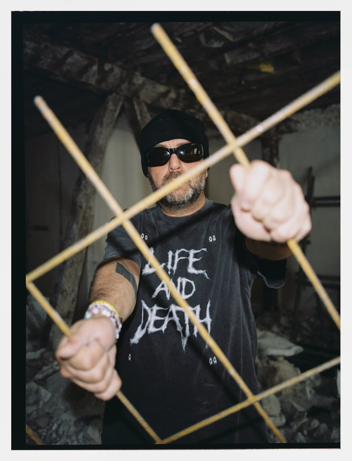 A bearded man in a beanie and sunglasses grips crossed bamboo poles in a derelict room, wearing a "Life and Death" T-shirt.
