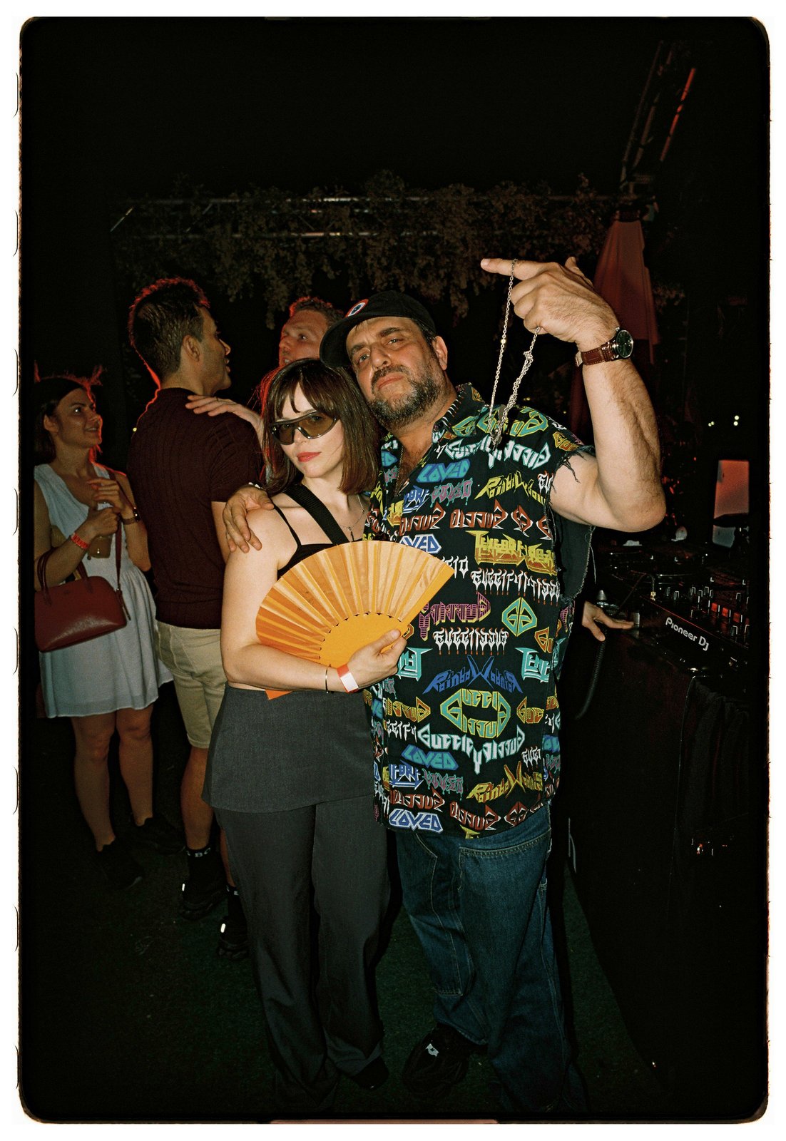 Night flash of a bearded man in a printed shirt hugging a woman in sunglasses holding an orange folding fan, DJ gear behind.