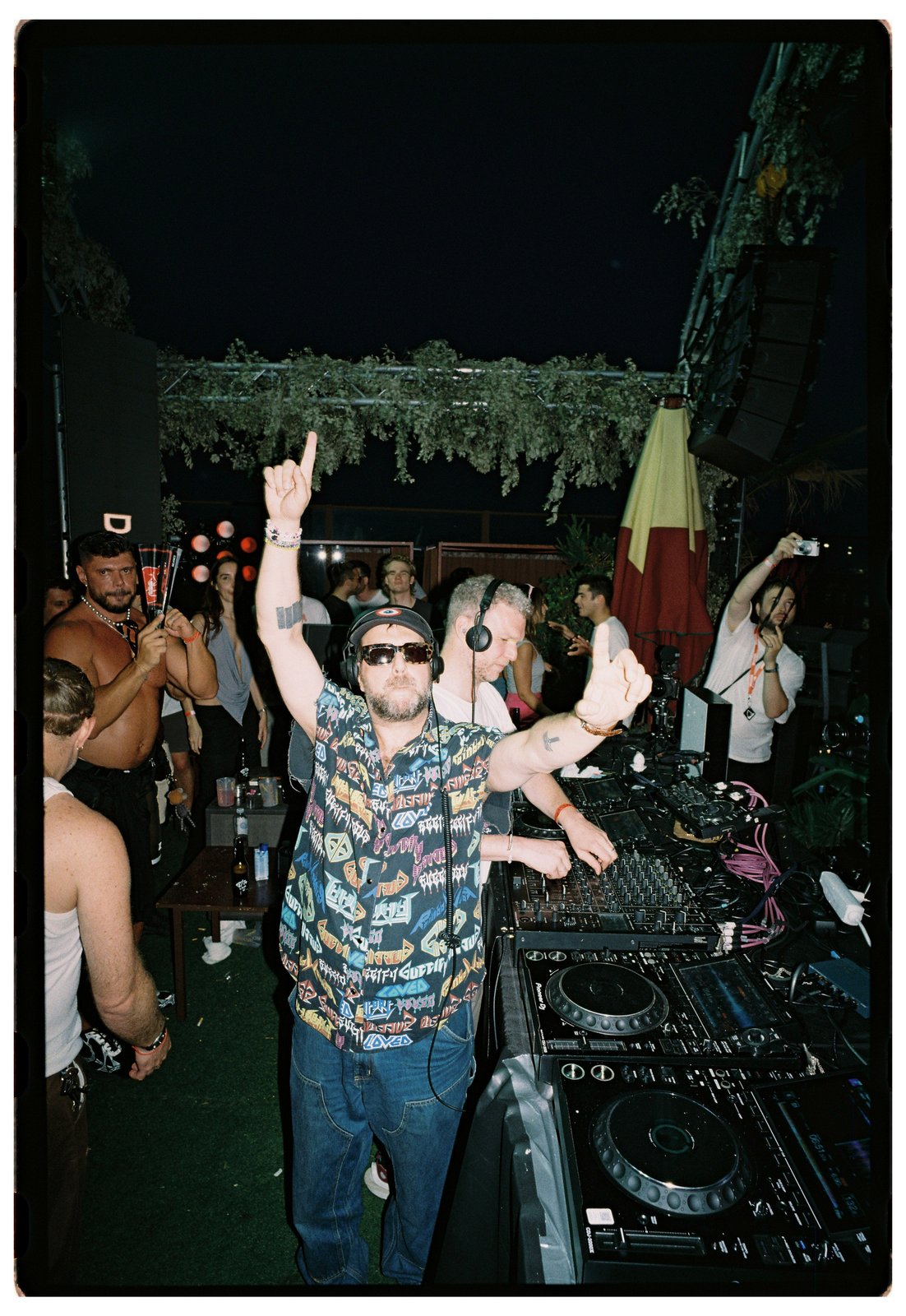 A man in headphones and a graphic-print shirt DJs at the decks with one arm raised, crowd around at a night party.