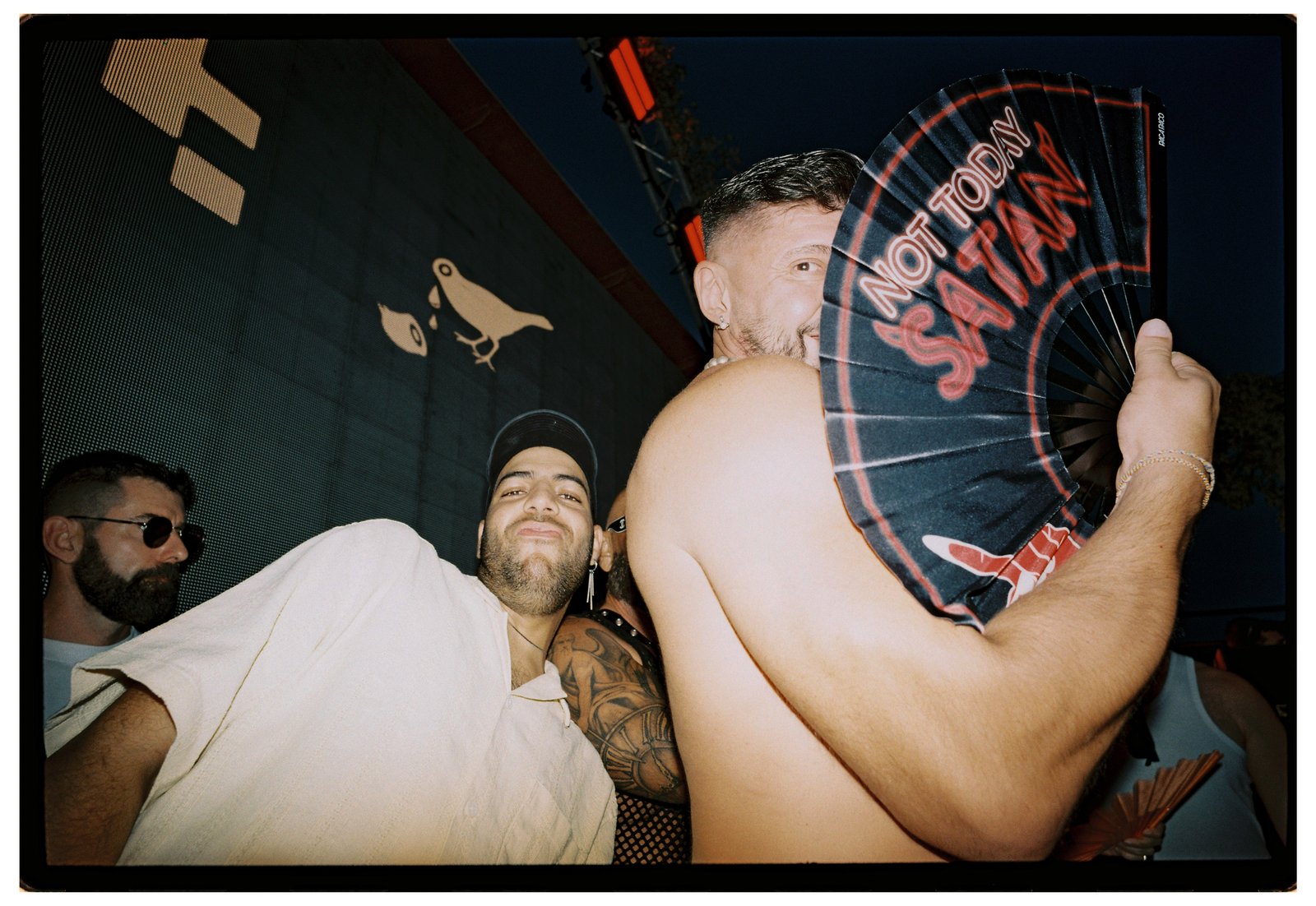 Low wide-angle night flash of two men close together, one holding a black hand fan lettered in red.