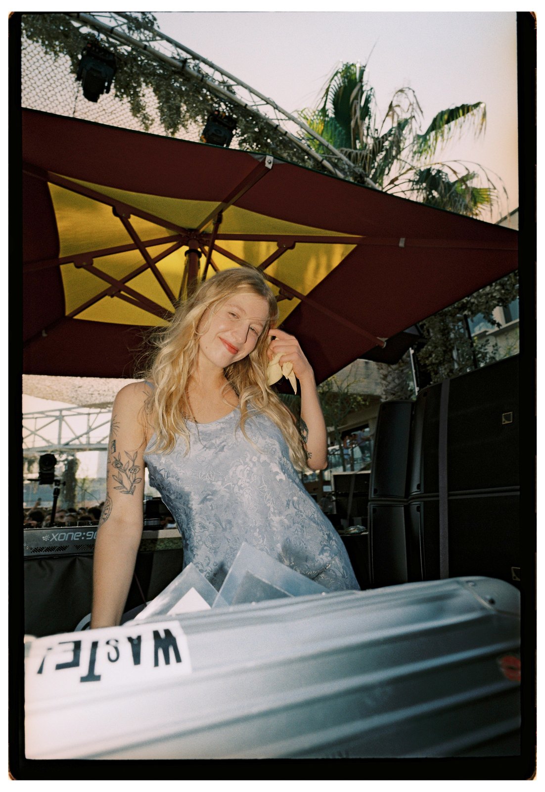 A smiling blonde woman in a pale-blue lace dress stands under a burgundy umbrella in daylight, palms and flight cases nearby.